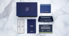 A chic flat lay showcases the limited-edition COMING SOON Jacques Marie Mage 1991 luxury eyewear box, glasses case, booklet, certificate, and documents—all in sleek navy blue designs on a marble surface.