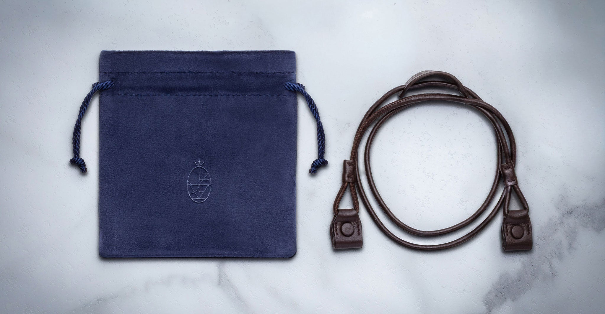 The Jacques Marie Mage 1992, featuring a blue suede drawstring pouch with embossed logo and a coiled dark brown leather strap with clasps, is displayed on a light marble surface.