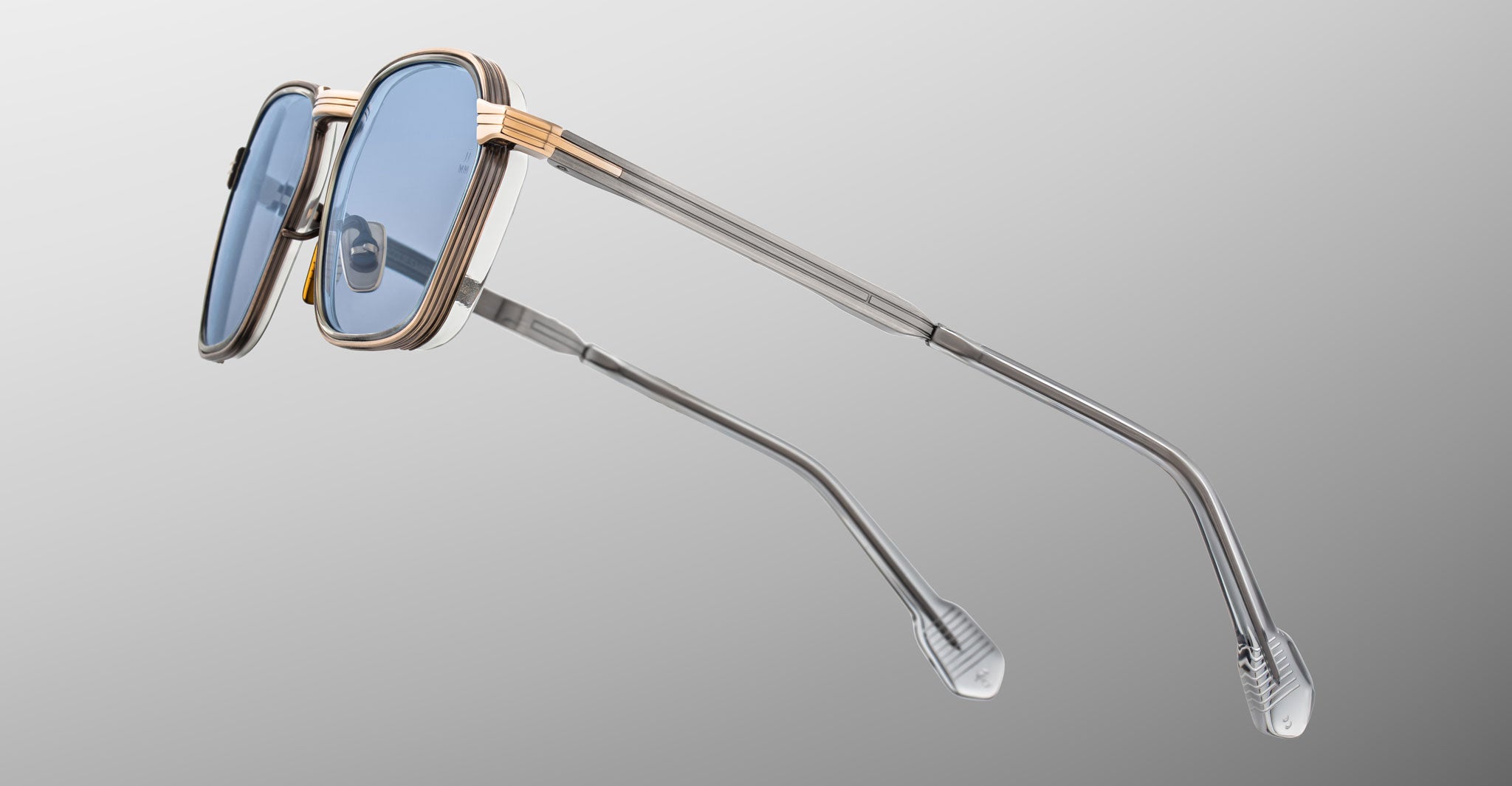 The Jacques Marie Mage Beckett sunglasses, handcrafted in Japan, feature blue-tinted lenses, a silver and gold dual-tone metal frame, thin temples with clear tips, and are shown against a light gray gradient background.