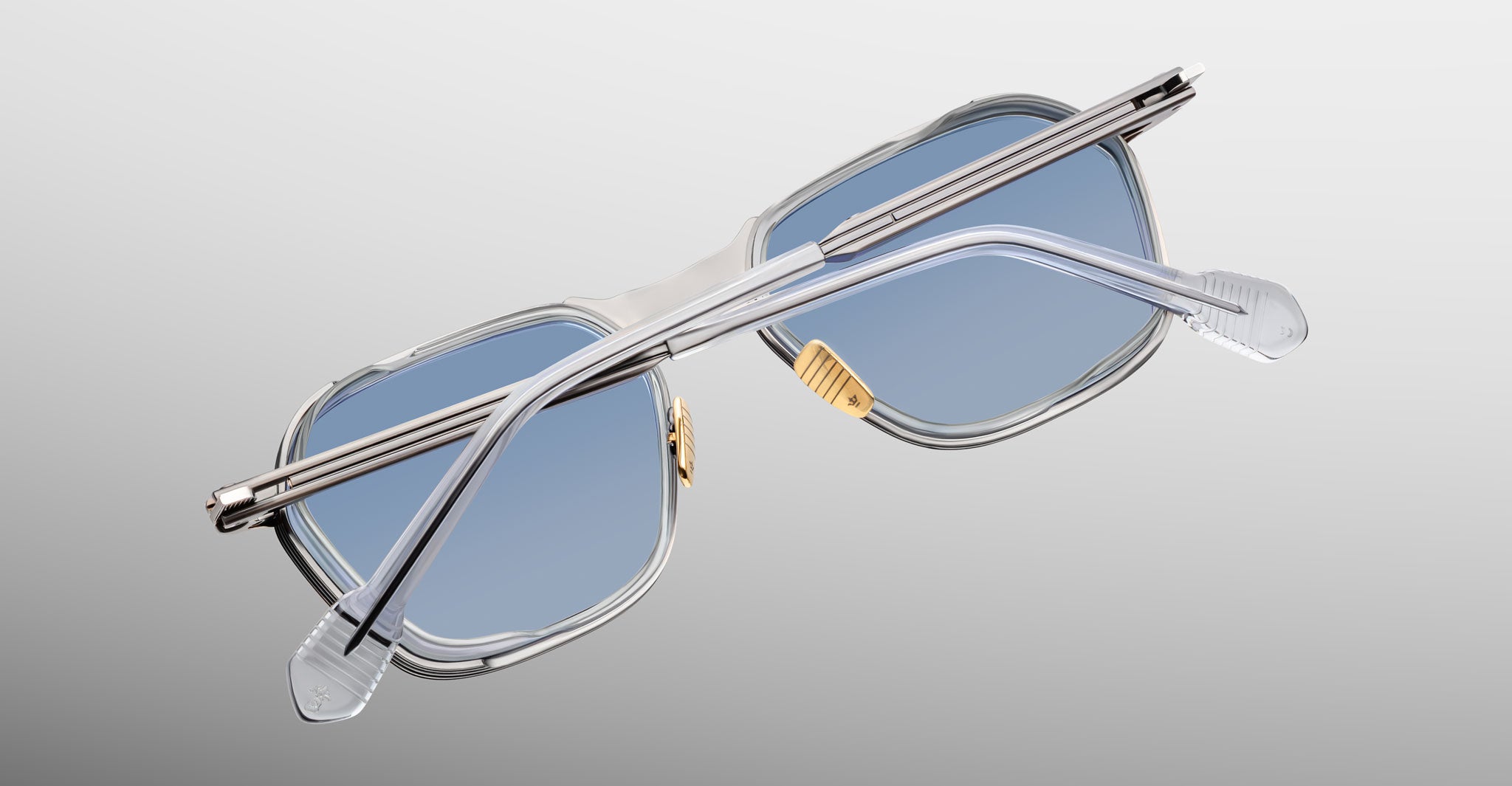A pair of Jacques Marie Mage Beckett sunglasses with blue-tinted lenses and clear rectangular frames, handcrafted in Japan, displayed from the back on a light gray gradient background.