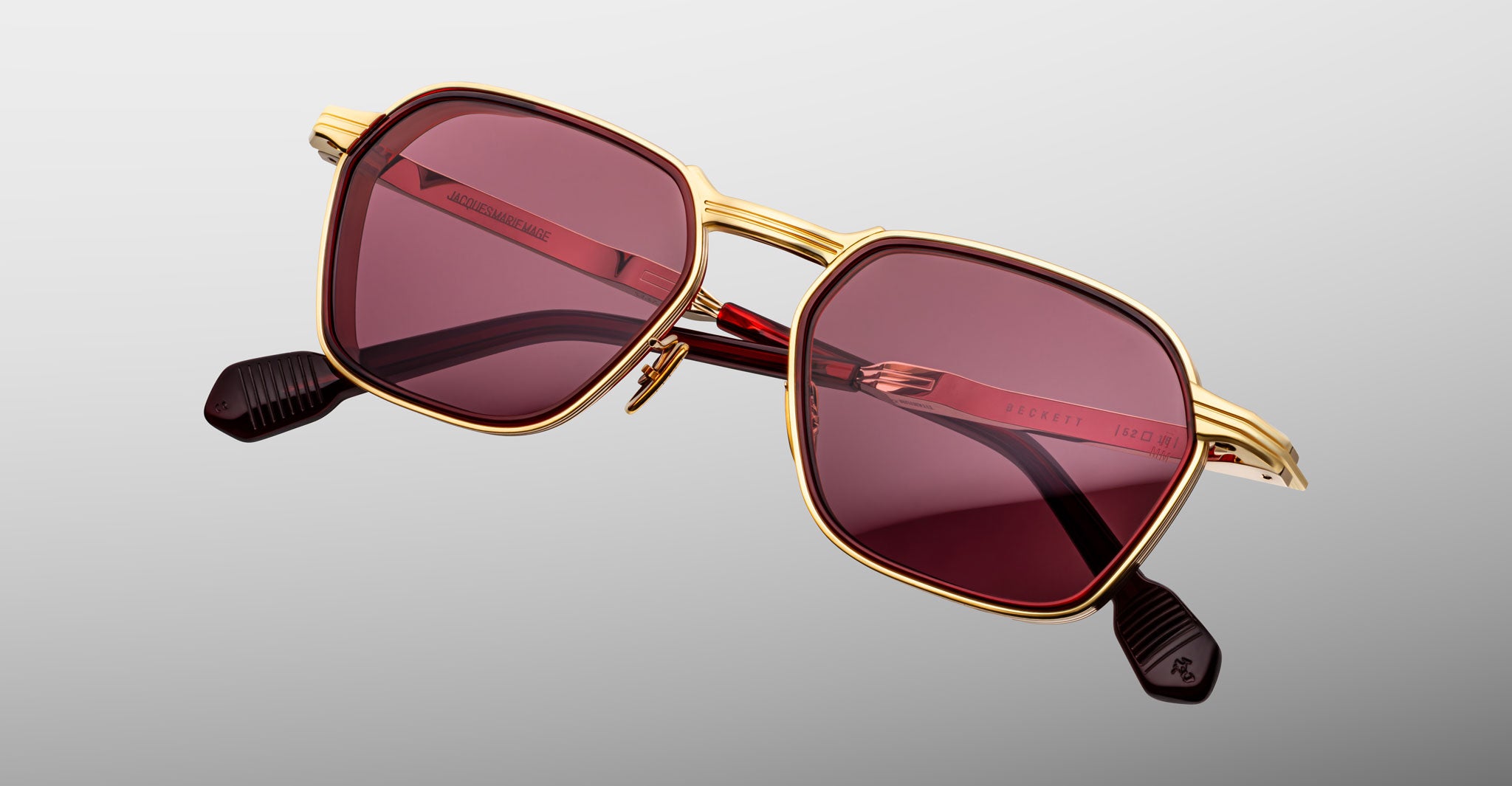 The Jacques Marie Mage Beckett sunglasses feature a gold frame, dark red lenses, and black temple tips. This limited-edition pair showcases a modern geometric shape with visible detailing on the arms and bridge, set against a light gray background.