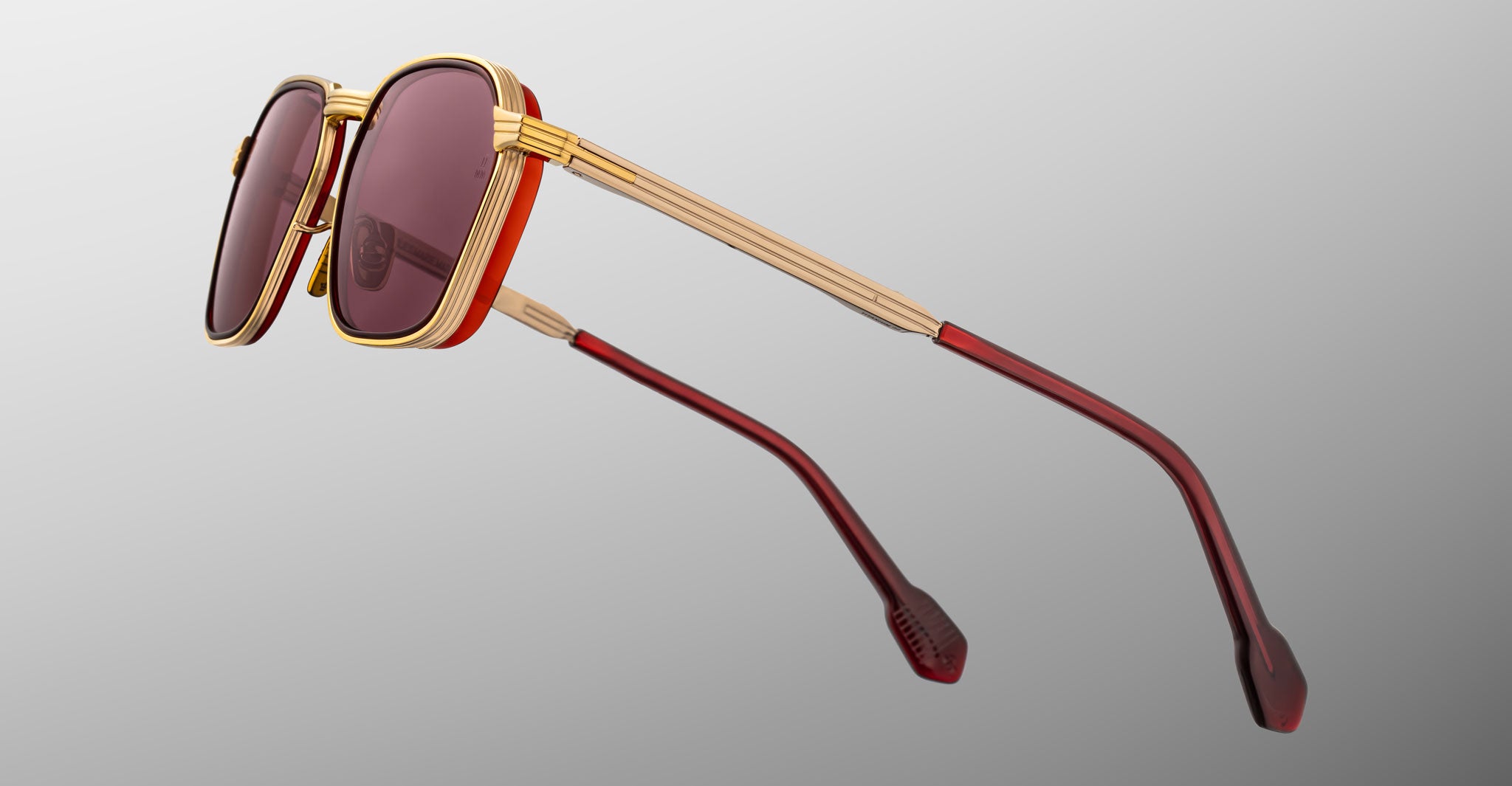 The Jacques Marie Mage Beckett sunglasses feature gold metal frames with red accents and dark red lenses. Shown from a side angle, this limited-edition piece stands out against a light gray background.