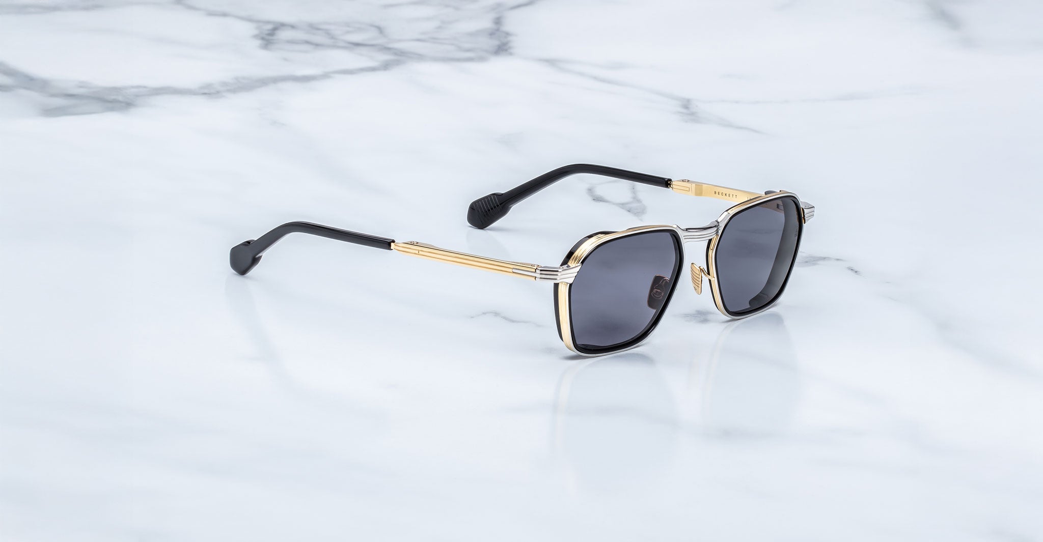A pair of Jacques Marie Mage Beckett sunglasses with gold and silver metal frames and black arms, handcrafted in Japan, rests on a smooth, white marble surface.