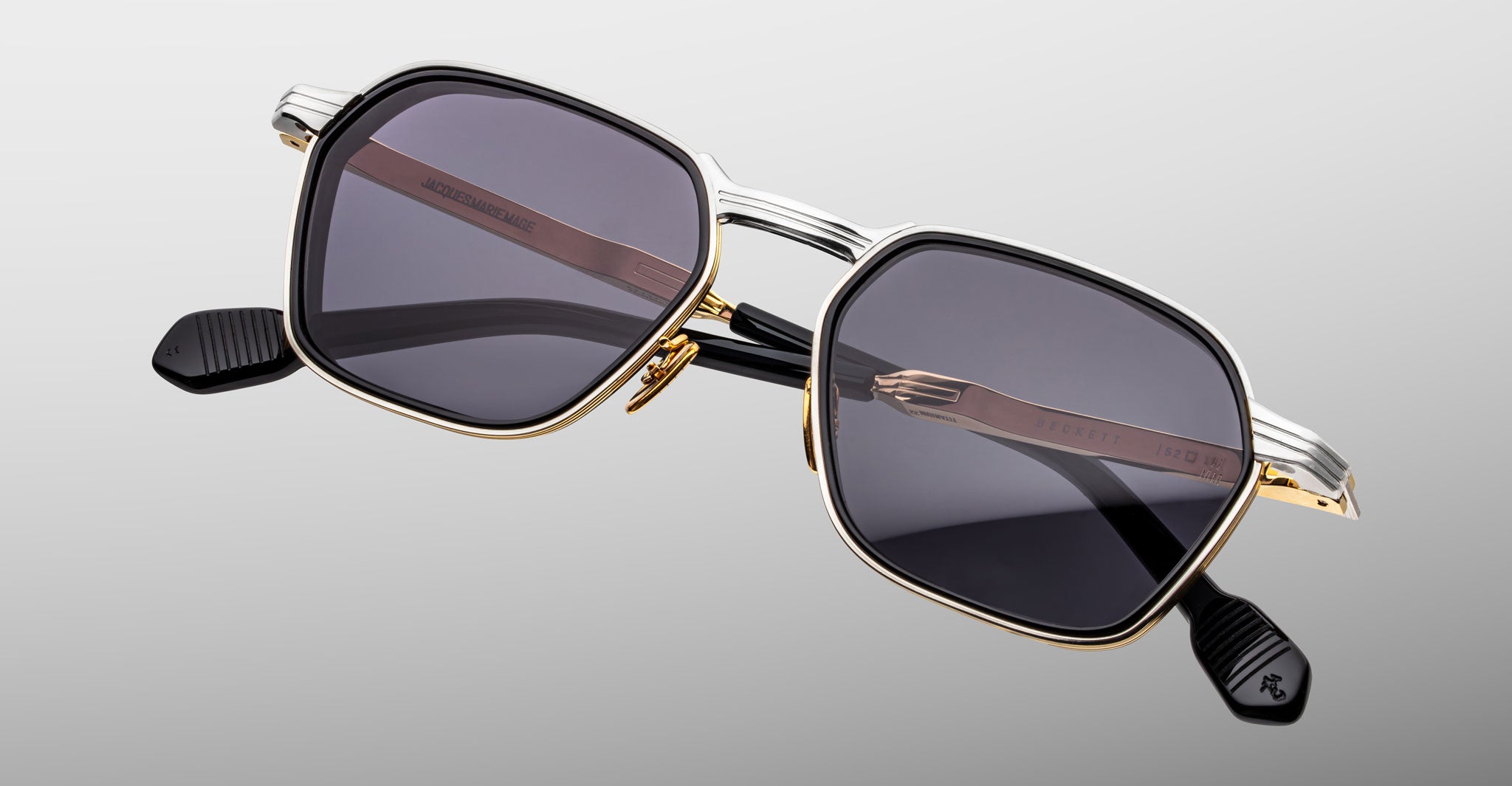 The Jacques Marie Mage Beckett sunglasses, handcrafted in Japan, feature gold and silver square metal frames, black arms, and dark tinted lenses displayed on a light gray background.