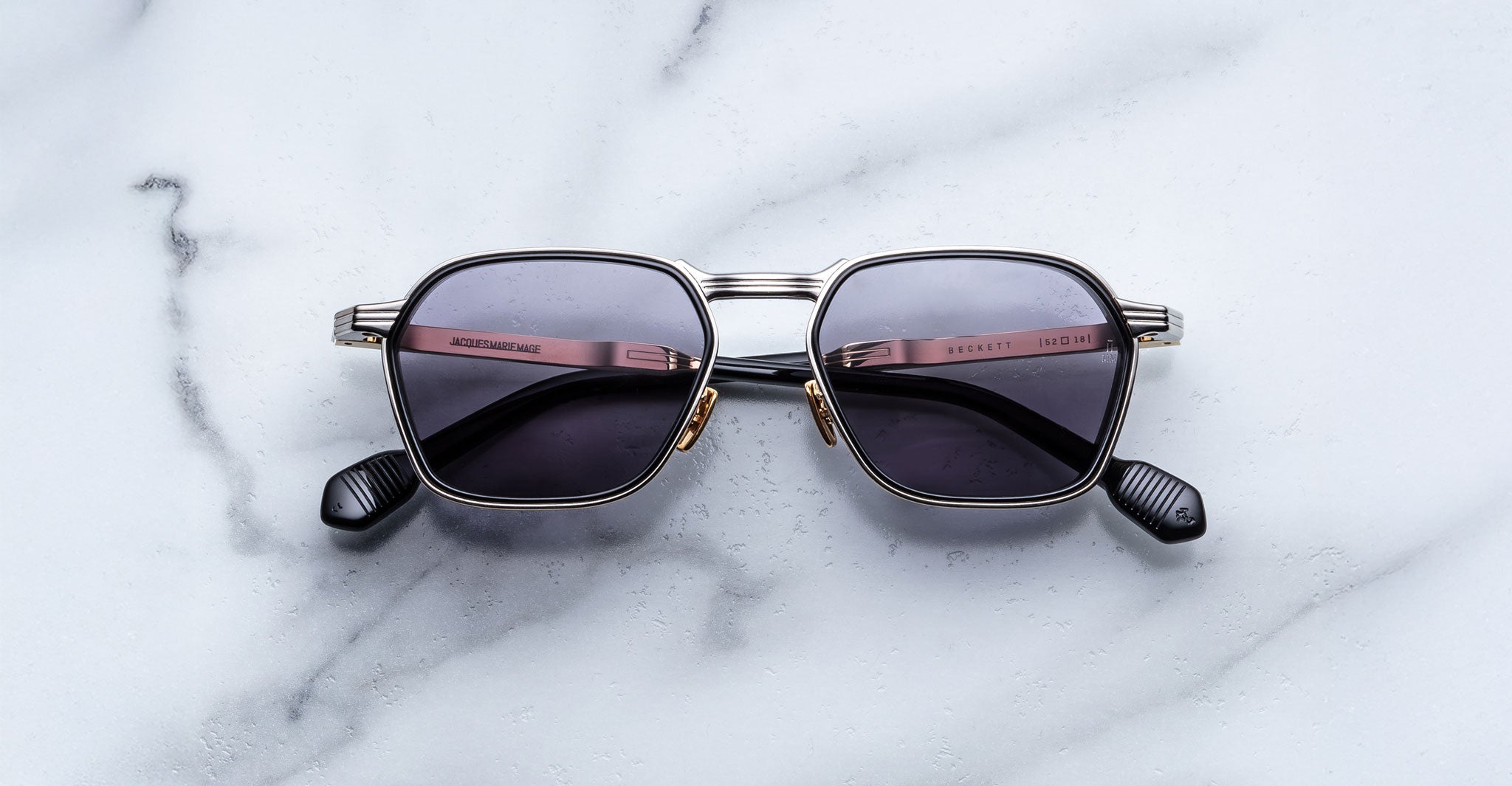A pair of Jacques Marie Mage Beckett sunglasses with square-shaped silver frames and dark lenses rests on white marble. This limited-edition frame features closed arms with black tips, reflecting exceptional style and craftsmanship.