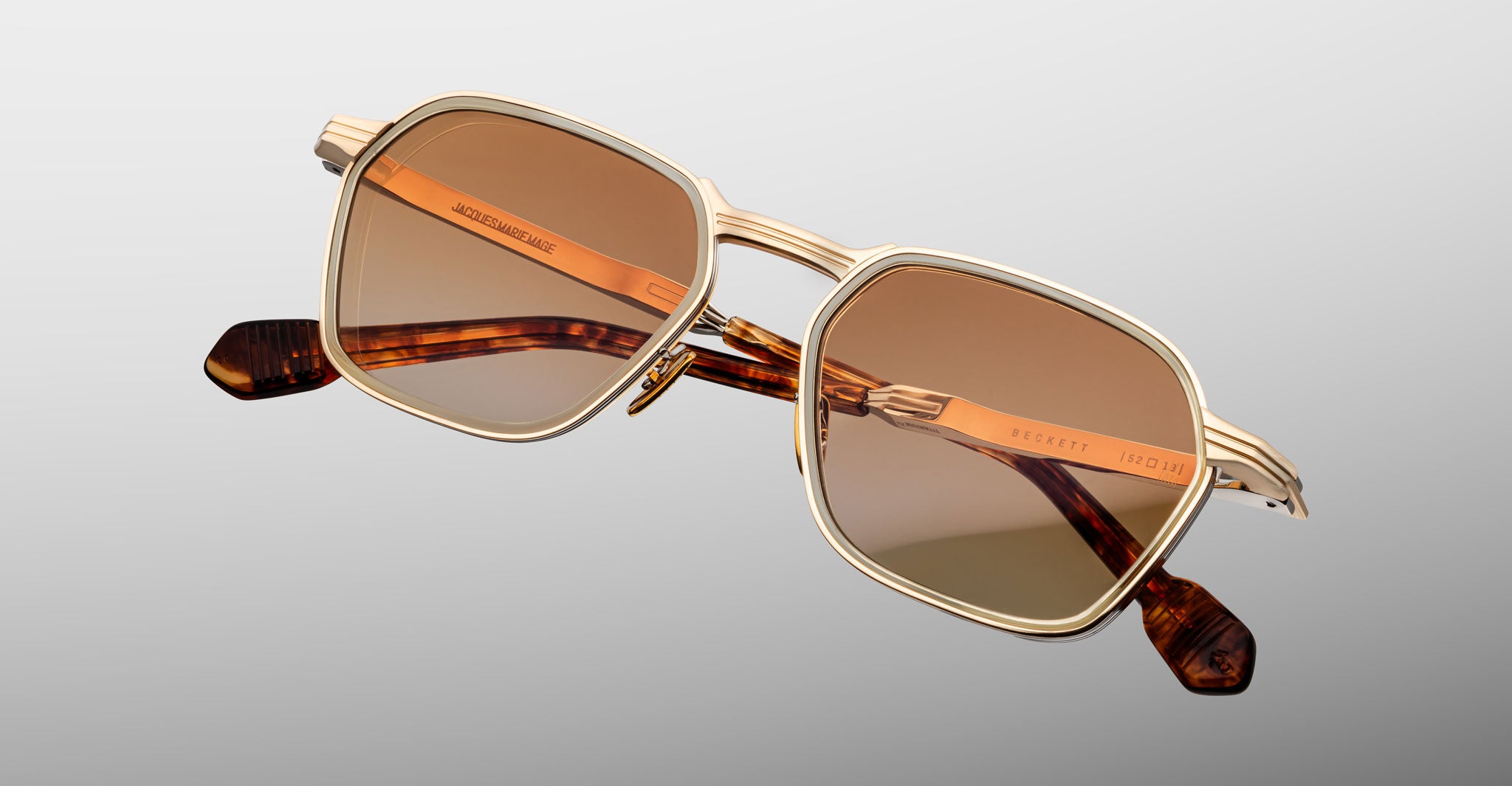 The Jacques Marie Mage Beckett sunglasses feature gold metal frames, square brown-tinted lenses, and tortoiseshell-patterned arms—this limited-edition pair is displayed against a light gray background.