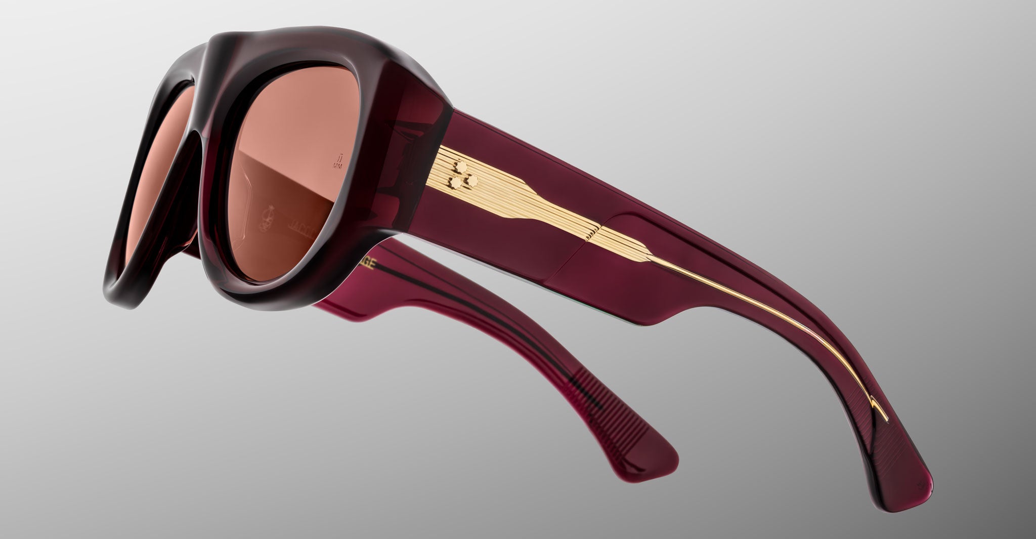 Close-up of Jacques Marie Mage Bel Air sunglasses featuring burgundy thick frames, brown-tinted lenses, and gold detailing on the arms, set against a plain gradient background.