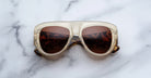 A pair of Jacques Marie Mage Bel Air sunglasses featuring oversized translucent beige handcrafted acetate frames and brown-tinted lenses sits on a white marble surface.