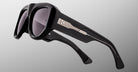A close-up of the Jacques Marie Mage Bel Air sunglasses, showcasing thick black glossy frames, dark lenses, and temples adorned with a silver metallic accent featuring three studs and a linear design against a plain gradient background.