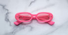 A pair of Jacques Marie Mage Besset bright pink oval sunglasses with tinted lenses is placed upside down on a white marble surface.