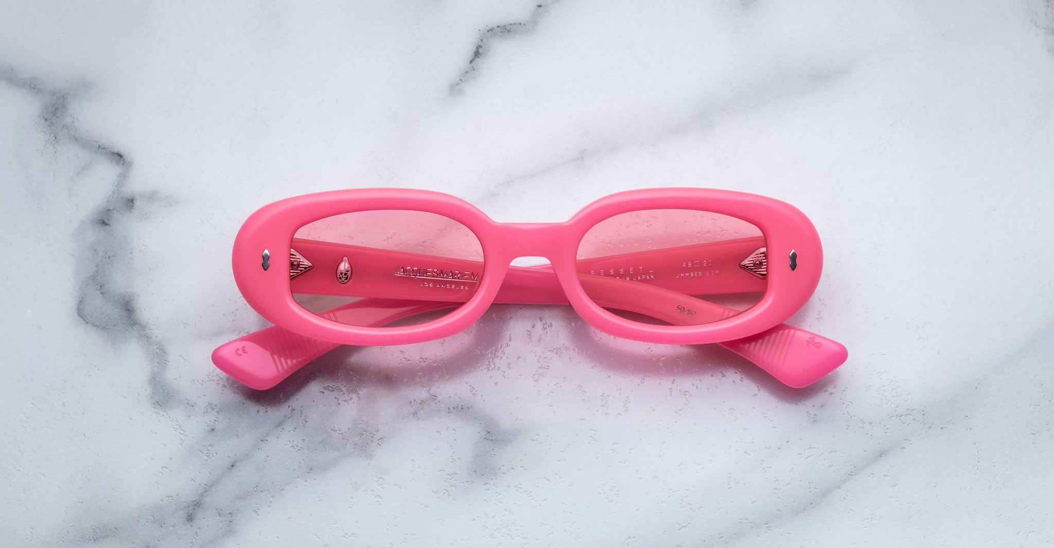 A pair of Jacques Marie Mage Besset bright pink oval sunglasses with tinted lenses is placed upside down on a white marble surface.