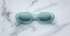 The Jacques Marie Mage Besset sunglasses, light blue and oval-shaped with tinted lenses, rest on a white marble surface with their arms slightly open and visible.