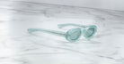 The Jacques Marie Mage Besset sunglasses, featuring thick transparent light blue frames, are displayed at an angle on a white marble surface with gray veining.