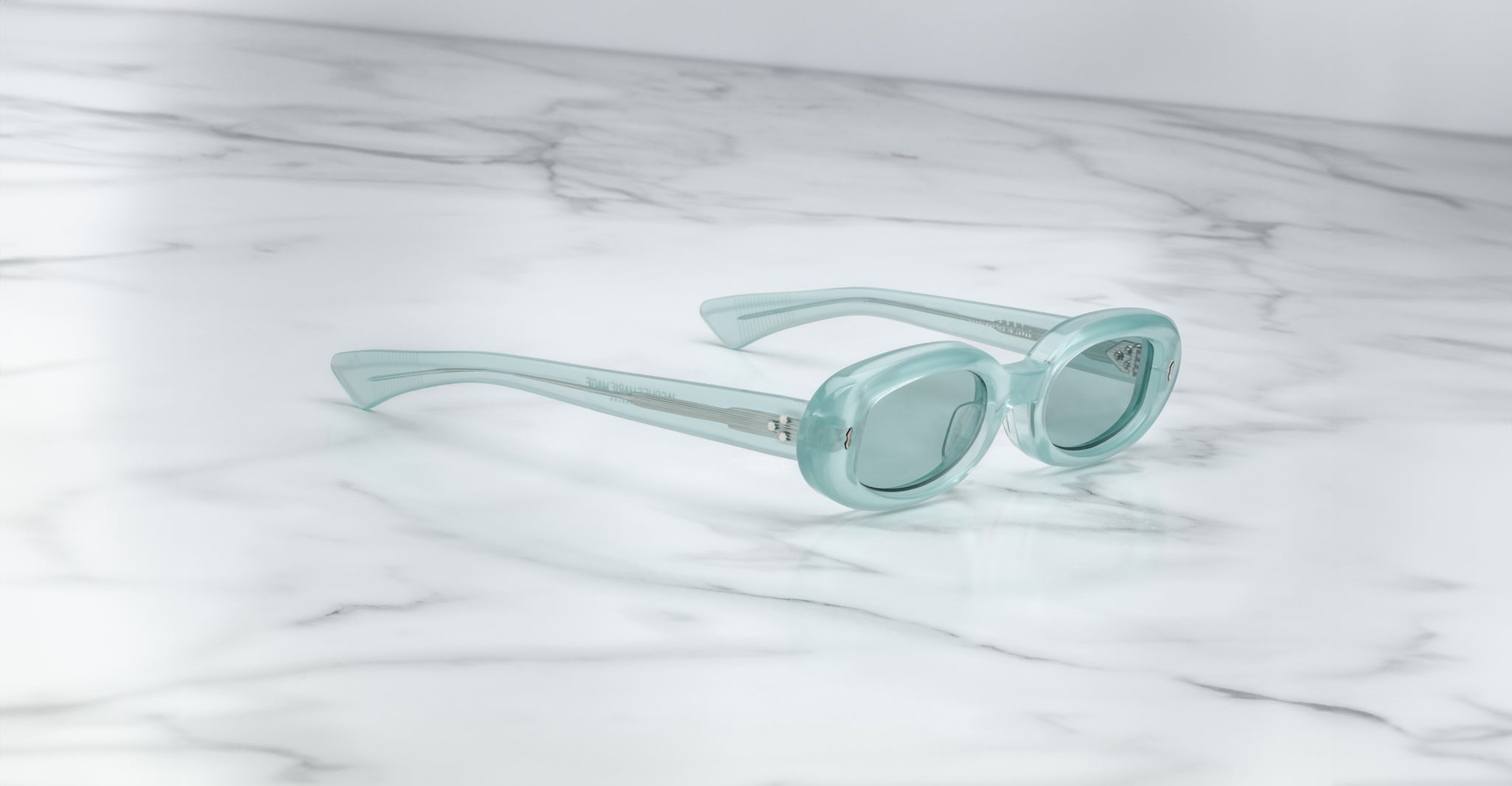The Jacques Marie Mage Besset sunglasses, featuring thick transparent light blue frames, are displayed at an angle on a white marble surface with gray veining.