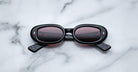 A pair of Jacques Marie Mage Besset black oval sunglasses with dark lenses rests face-up on a white marble surface with gray veining, their arms crossed in an X shape.