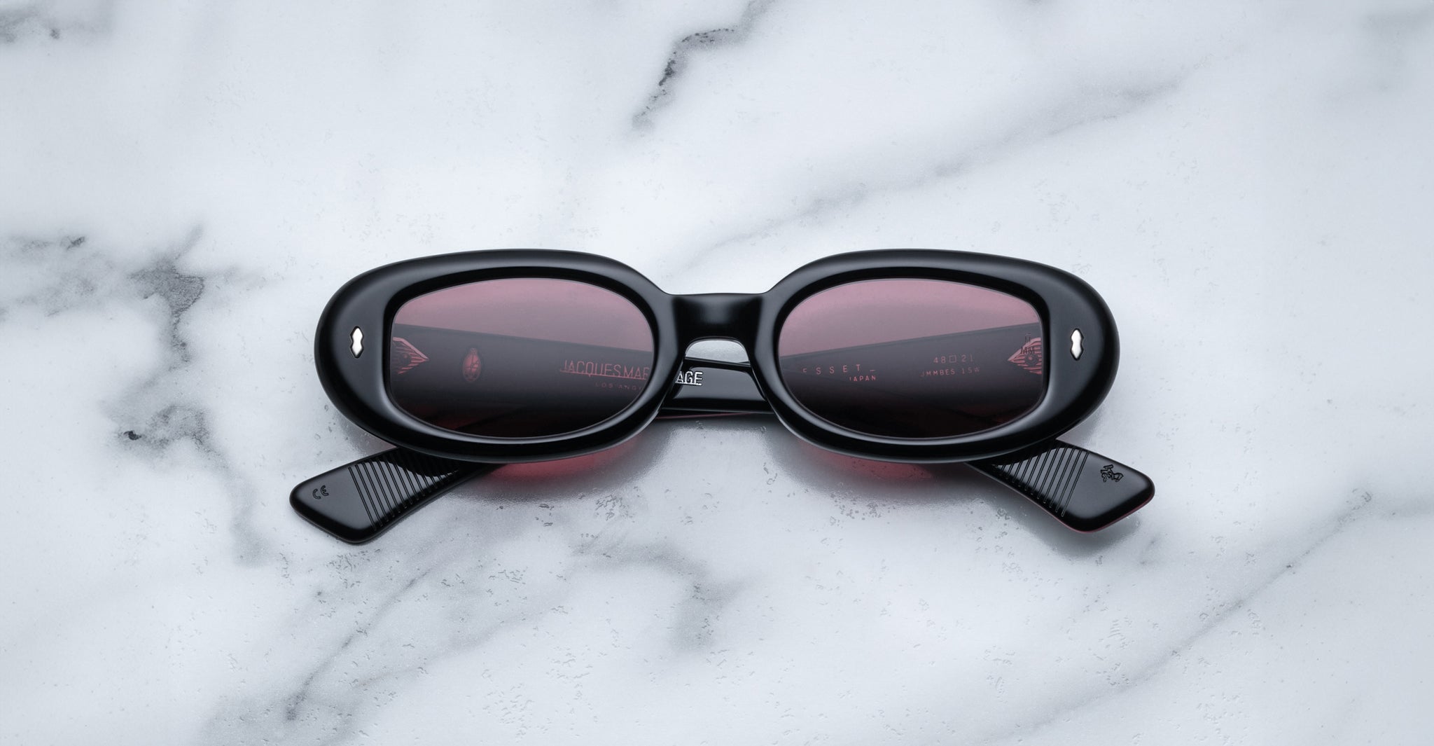 A pair of Jacques Marie Mage Besset black oval sunglasses with dark lenses rests face-up on a white marble surface with gray veining, their arms crossed in an X shape.