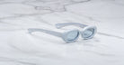 Jacques Marie Mage Besset sunglasses feature light blue, oval-shaped thick frames and lightly tinted lenses, elegantly displayed on a white marble surface.