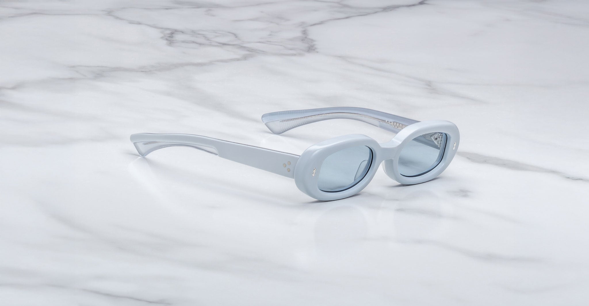 Jacques Marie Mage Besset sunglasses feature light blue, oval-shaped thick frames and lightly tinted lenses, elegantly displayed on a white marble surface.