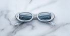 A pair of Jacques Marie Mage Besset white oval sunglasses with blue-tinted lenses rests on a white marble surface. The crossed temples and a metal railing reflected in the lenses add to their striking appearance.