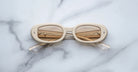 The Jacques Marie Mage Besset, a pair of cream-colored oval sunglasses with tinted lenses and folded arms, rests upside down on a white marble surface.
