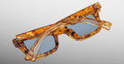 The Jacques Marie Mage Casablanca sunglasses, featuring a marbled brown tortoiseshell frame with blue lenses and silver accents on the front corners, are shown folded on a light grey surface.