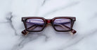 A pair of Jacques Marie Mage Casablanca square eyeglasses with dark red frames and clear lenses rests on a white marble surface, the partially folded arms and faint lens reflection visible.