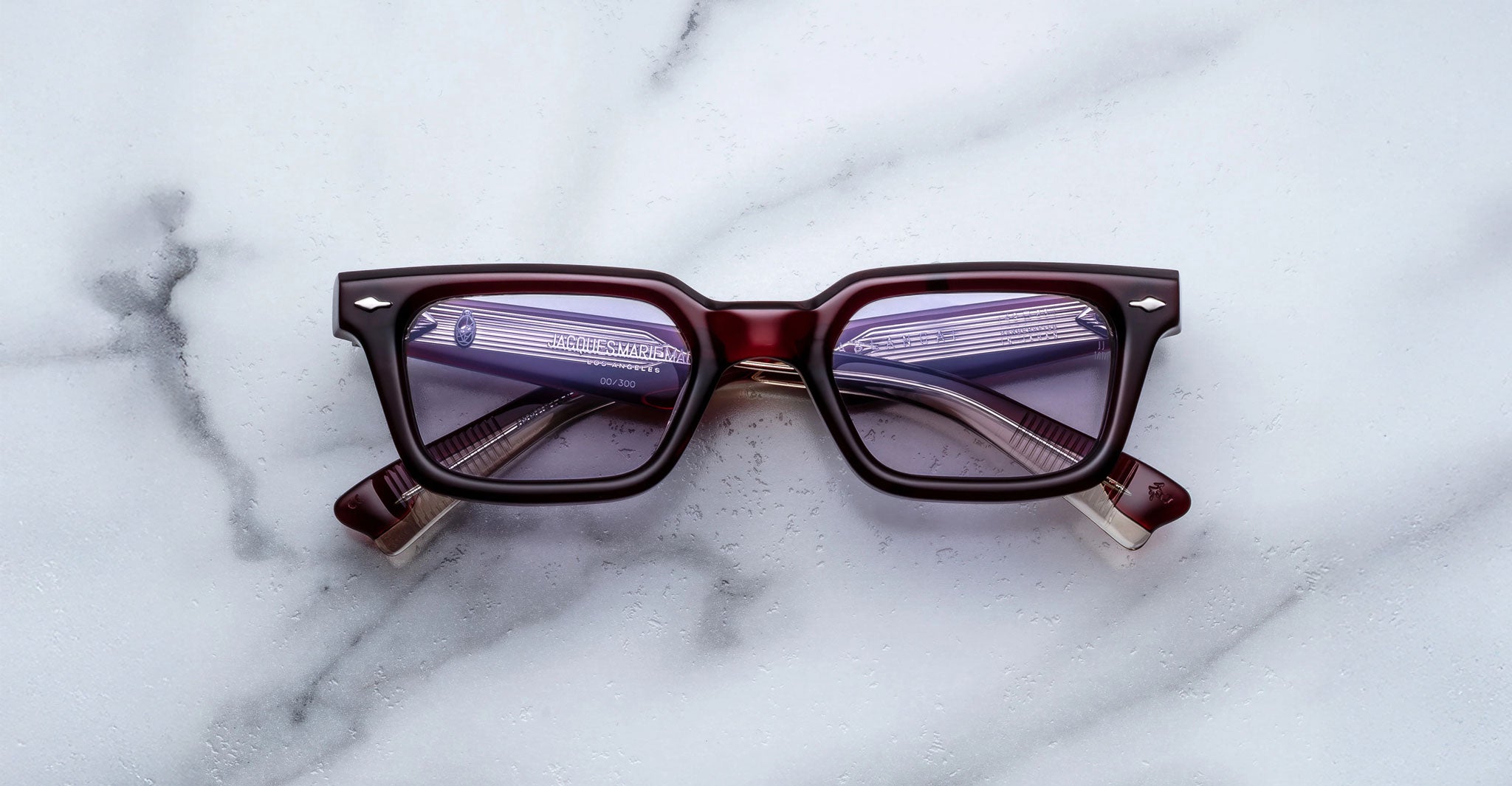 A pair of Jacques Marie Mage Casablanca square eyeglasses with dark red frames and clear lenses rests on a white marble surface, the partially folded arms and faint lens reflection visible.