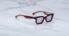 The Jacques Marie Mage Casablanca eyeglasses feature a rectangular, thick frame in dark red with clear accents, tinted lenses, and gold-toned arm details, displayed on a white marble surface.