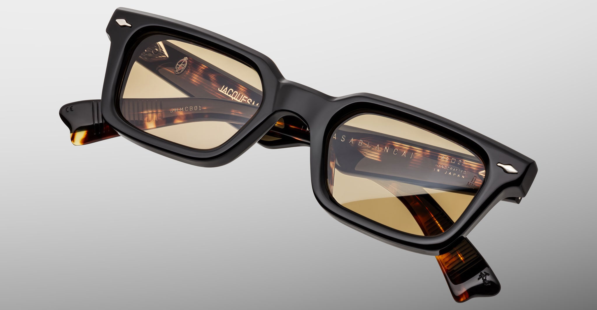 The Jacques Marie Mage Casablanca features rectangular black acetate frames with thick edges and light brown tinted lenses, showcased at an angle on a plain light gray background.