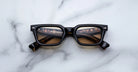 The Jacques Marie Mage Casablanca eyeglasses, featuring a black rectangular frame and brown tinted lenses, rest on a white marble surface with lights and text reflected in the lenses.