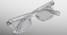 The Jacques Marie Mage Casablanca is a pair of clear, rectangular eyeglasses with thick transparent frames, folded and placed on a flat surface against a gradient gray background.
