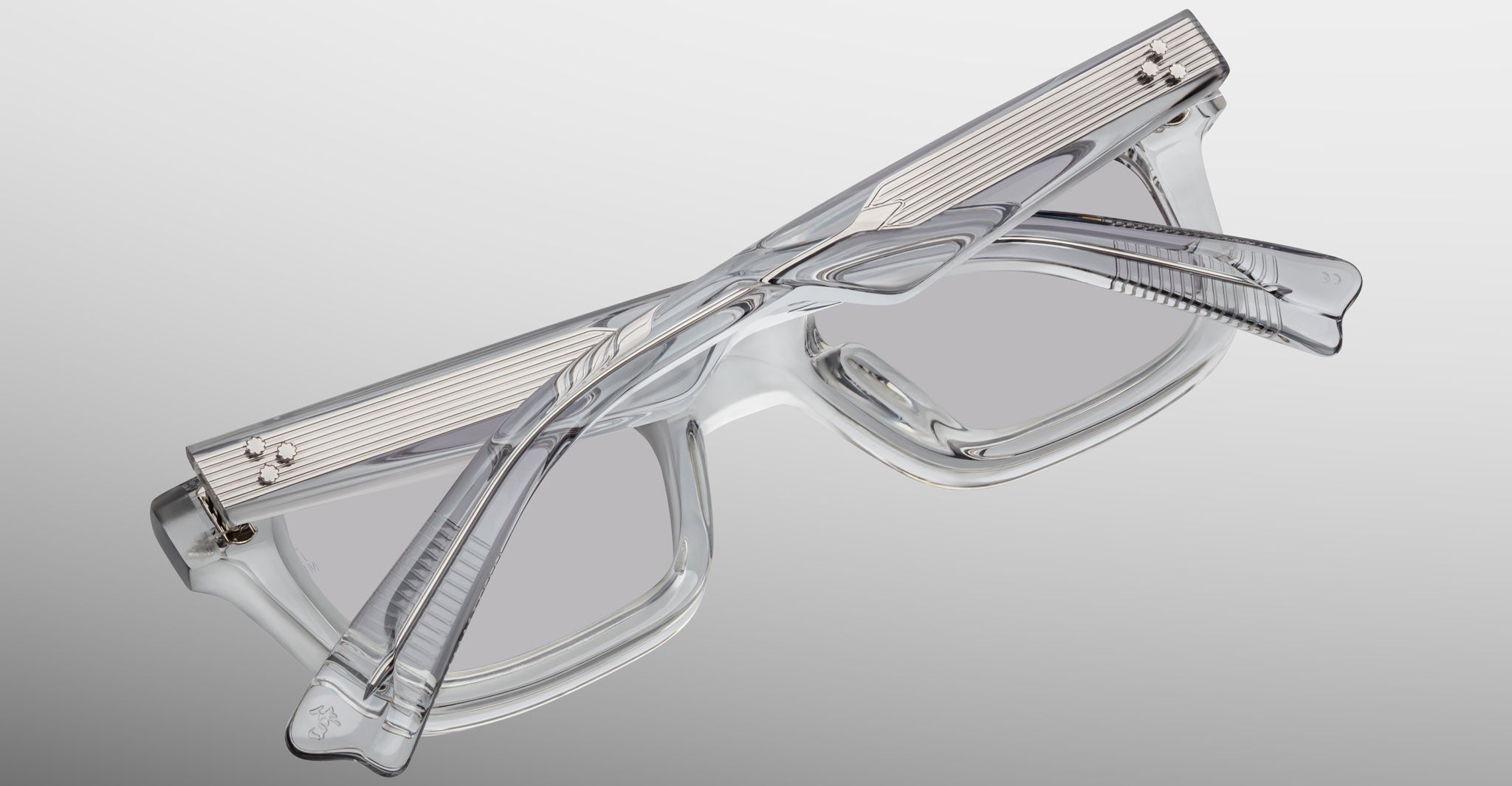 The Jacques Marie Mage Casablanca is a pair of clear, rectangular eyeglasses with thick transparent frames, folded and placed on a flat surface against a gradient gray background.