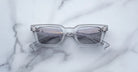 A pair of Jacques Marie Mage Casablanca rectangular sunglasses with thick translucent gray frames and tinted lenses rests on a white marble surface.
