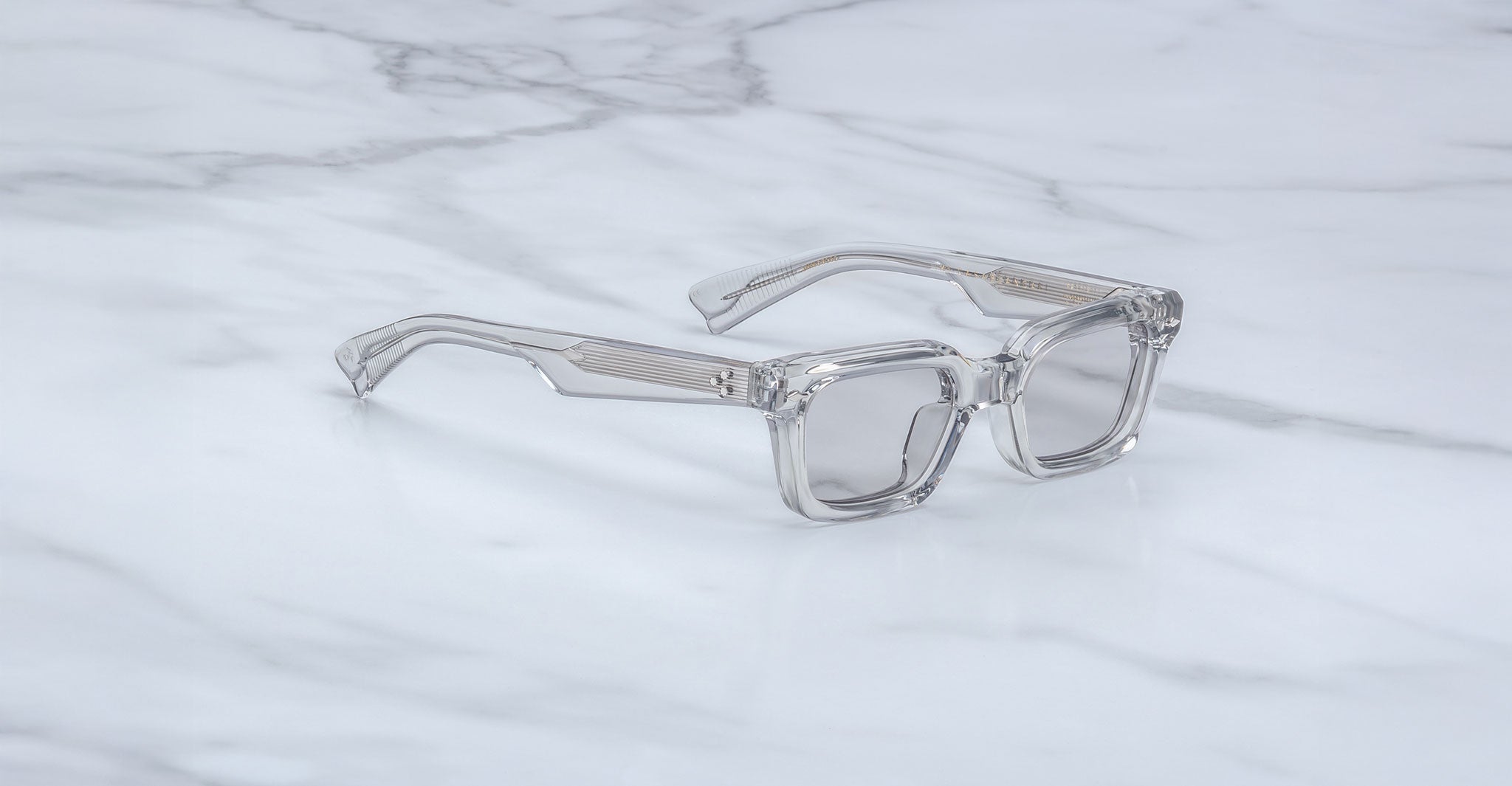 The Jacques Marie Mage Casablanca, a pair of clear, rectangular eyeglasses with thick frames, rests on a white marble surface streaked with gray veins.