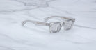 The Jacques Marie Mage Casablanca, a pair of clear, rectangular eyeglasses with thick frames, rests on a white marble surface streaked with gray veins.