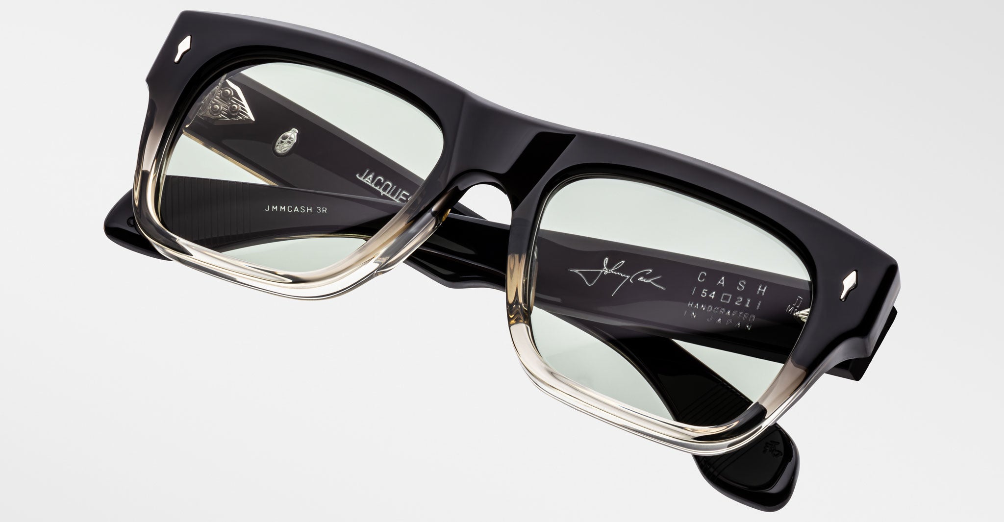 A close-up shows the Jacques Marie Mage Cash rectangular black and clear acetate eyeglasses with green-tinted lenses, highlighting engraved logos, text, and a signature on the inside of the temples.