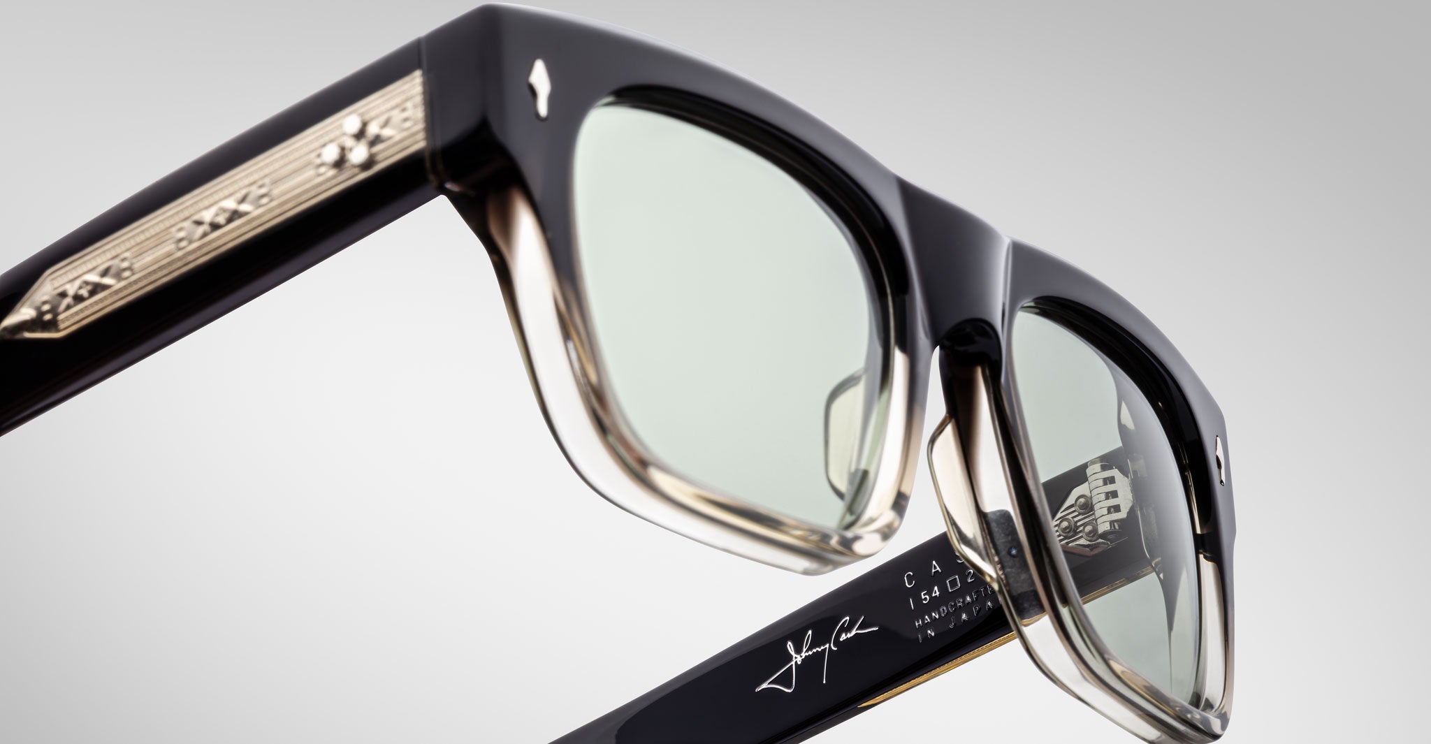 Close-up of Jacques Marie Mage Cash black and gold eyeglasses with engraved arm detailing, transparent lenses, and branded signature on the inside of the right temple, set against a light gray background.