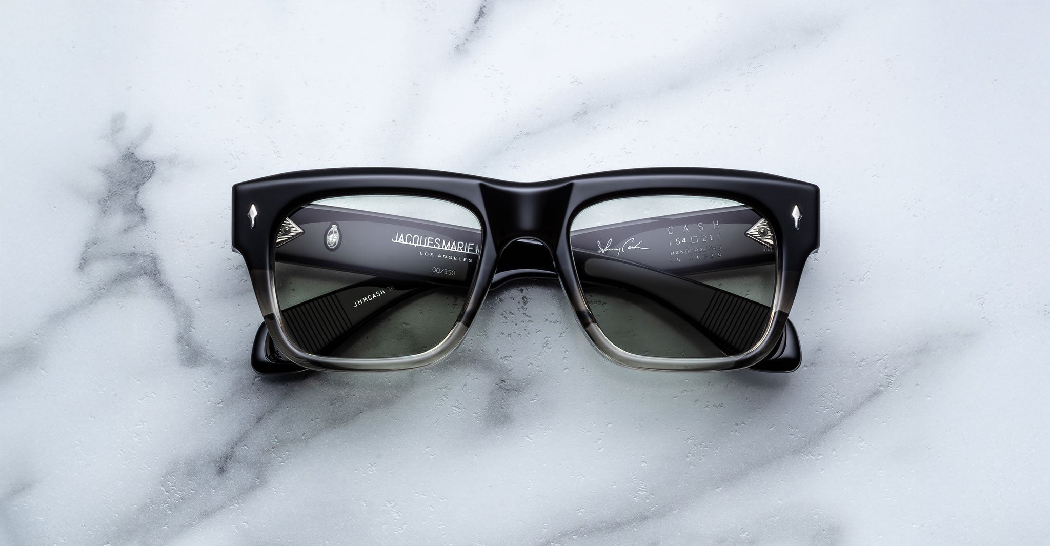 A pair of Jacques Marie Mage Cash black rectangular sunglasses with slightly translucent lenses rests on a white marble surface; the inner arms display branding and product details in white text.