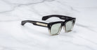 The Jacques Marie Mage Cash sunglasses, featuring a black square frame with metallic temple accents and lightly tinted lenses, are shown on a white marble surface.