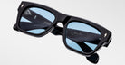 A pair of Jacques Marie Mage Cash black rectangular sunglasses with light blue tinted lenses is displayed against a plain background. The inner arms show white and gold branding, signature, and product details.