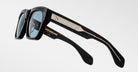 A pair of black Jacques Marie Mage Cash sunglasses with rectangular blue-tinted lenses, metallic arm details, and engraved text including the brand name in gold on the inner side.