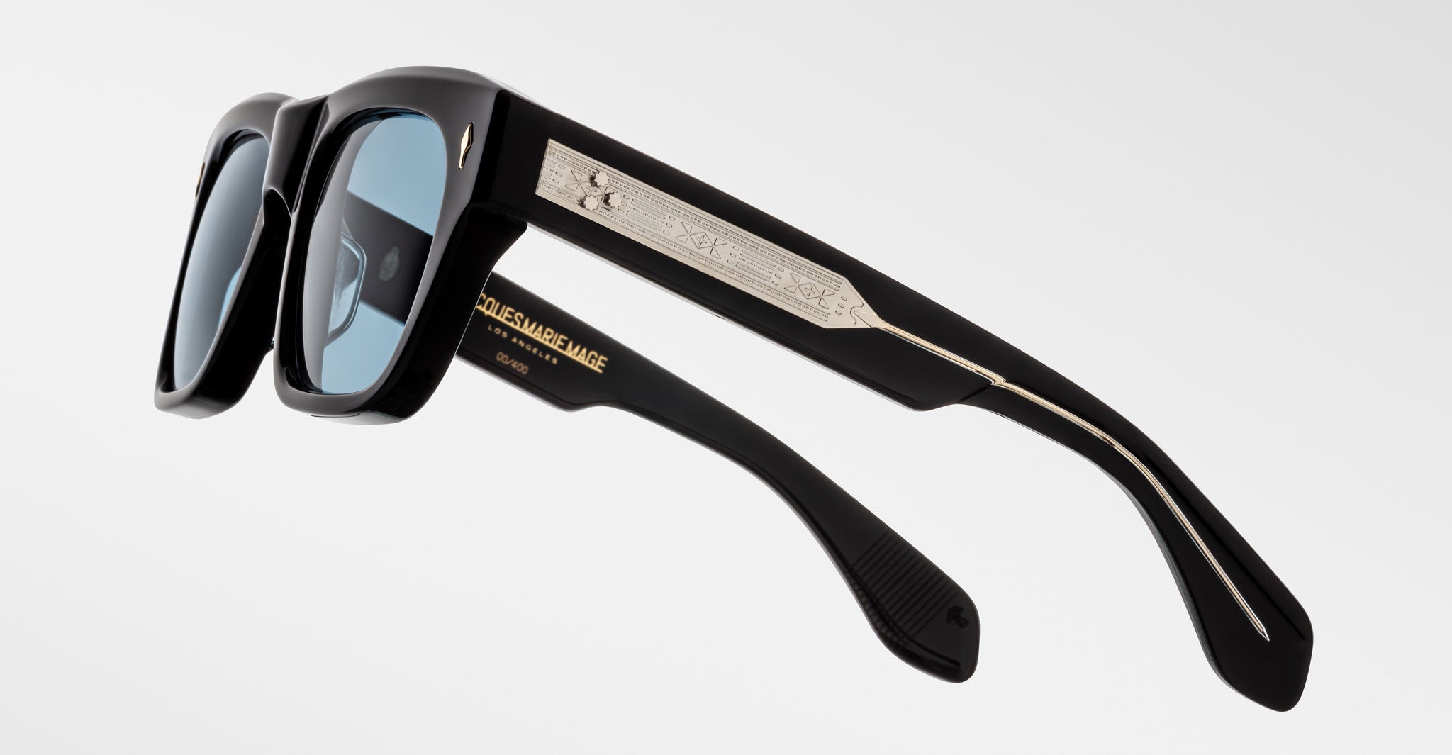 A pair of black Jacques Marie Mage Cash sunglasses with rectangular blue-tinted lenses, metallic arm details, and engraved text including the brand name in gold on the inner side.
