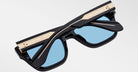 The Jacques Marie Mage Cash features black rectangular frames, light blue lenses, gold detailing on the top frame, and gold accents on the temples, shown against a plain white background.