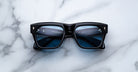 A pair of Jacques Marie Mage Cash black rectangular sunglasses with blue-tinted lenses rests on a white marble surface, with the partially visible arms seen through the lenses.