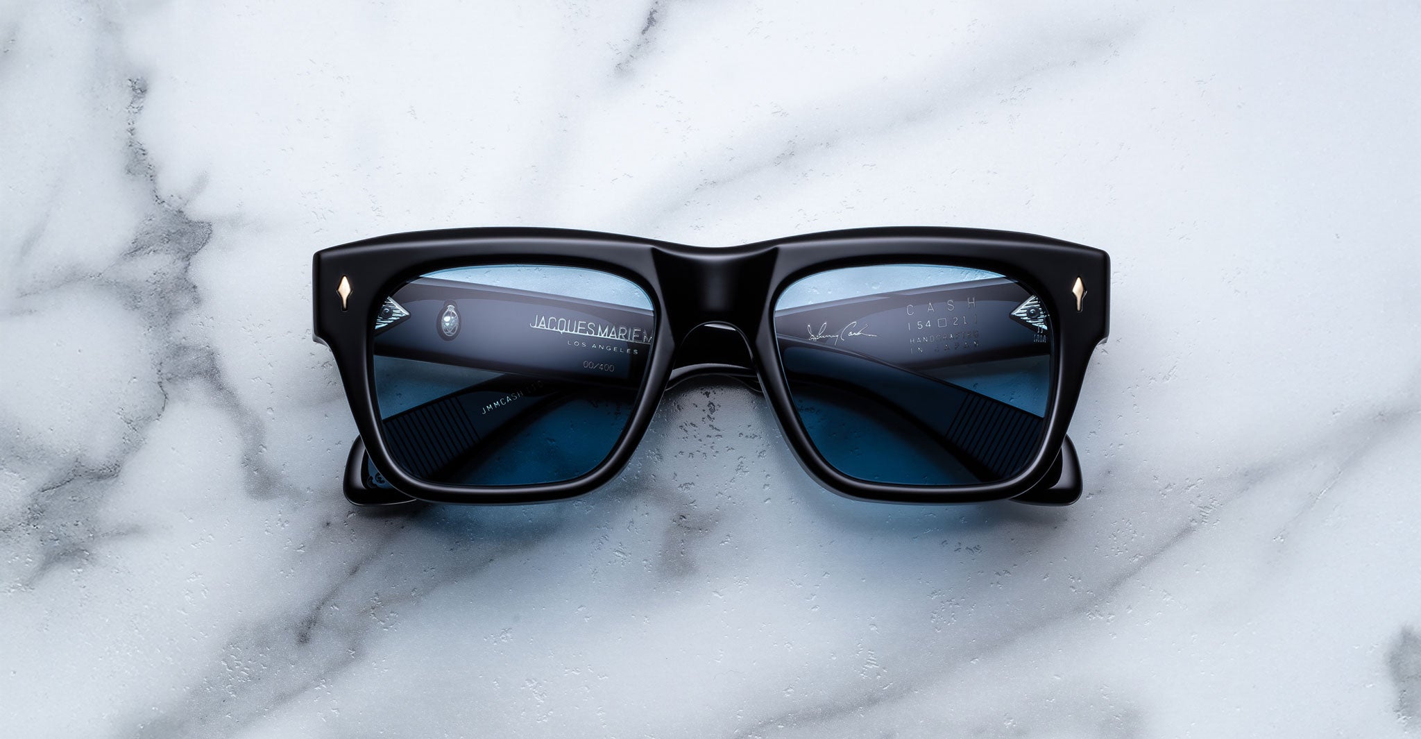 A pair of Jacques Marie Mage Cash black rectangular sunglasses with blue-tinted lenses rests on a white marble surface, with the partially visible arms seen through the lenses.