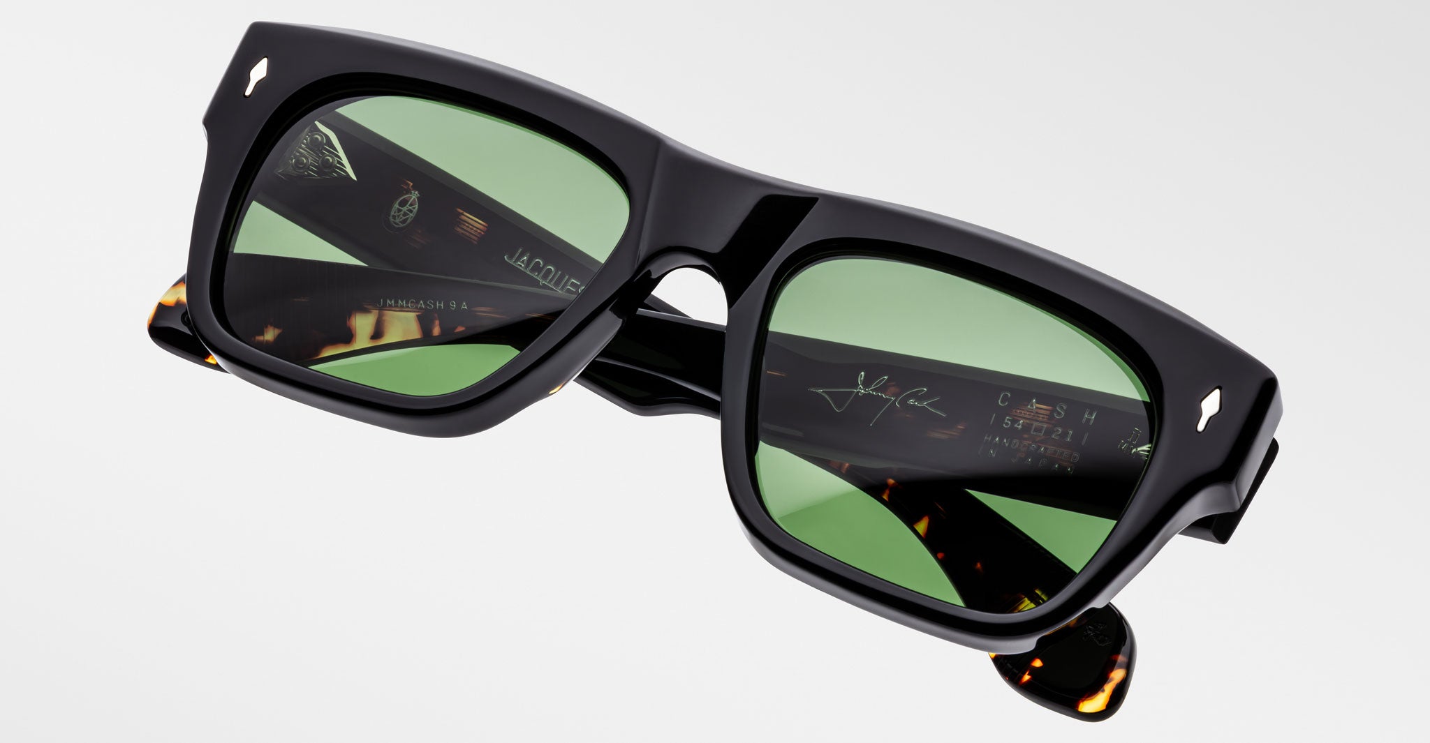 The Jacques Marie Mage Cash sunglasses feature black rectangular frames, green lenses, and tortoiseshell-detailed arms with branded and etched signatures on the inner sides, shown against a plain light background.