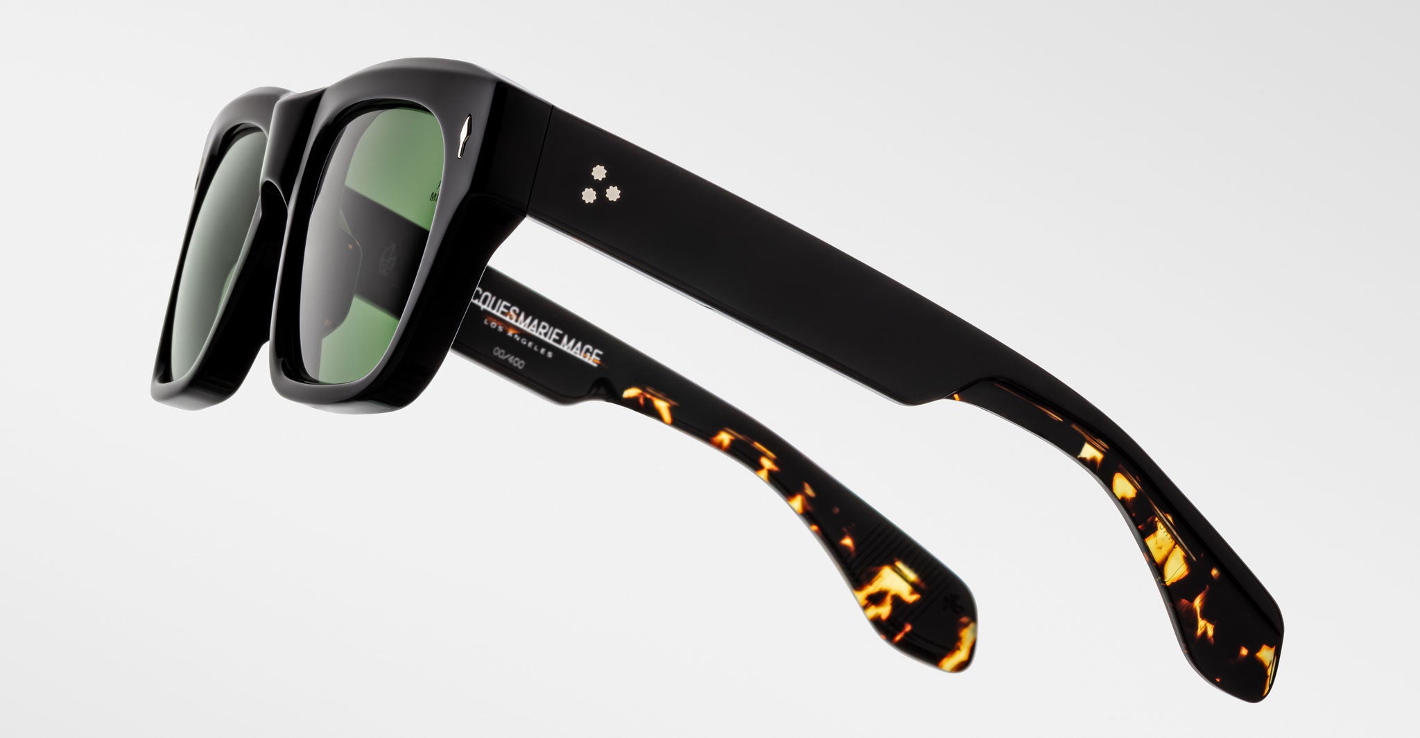 The Jacques Marie Mage Cash sunglasses feature thick black frames, green-tinted lenses, two small circular logos on one temple, and tortoiseshell detailing on the lower arms.