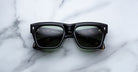 A pair of Jacques Marie Mage Cash black rectangular sunglasses with dark lenses rests on a white marble surface, reflecting dashboard instruments on the lenses.