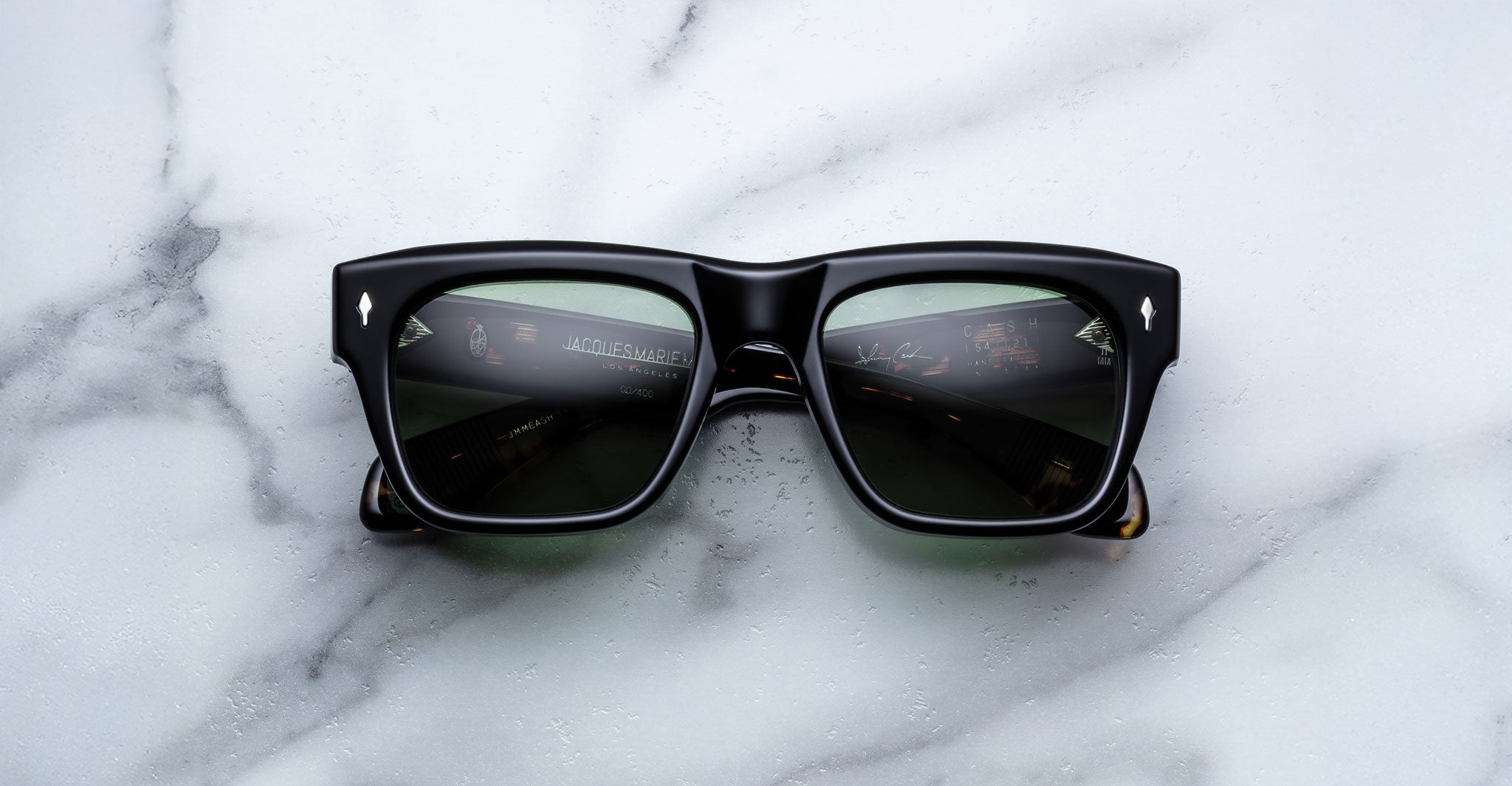 A pair of Jacques Marie Mage Cash black rectangular sunglasses with dark lenses rests on a white marble surface, reflecting dashboard instruments on the lenses.