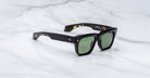 The Jacques Marie Mage Cash sunglasses feature black rectangular frames with green lenses, a slightly thick design, and rest on a white marble surface. The inner arms reveal a tortoiseshell pattern and illustrated detailing.